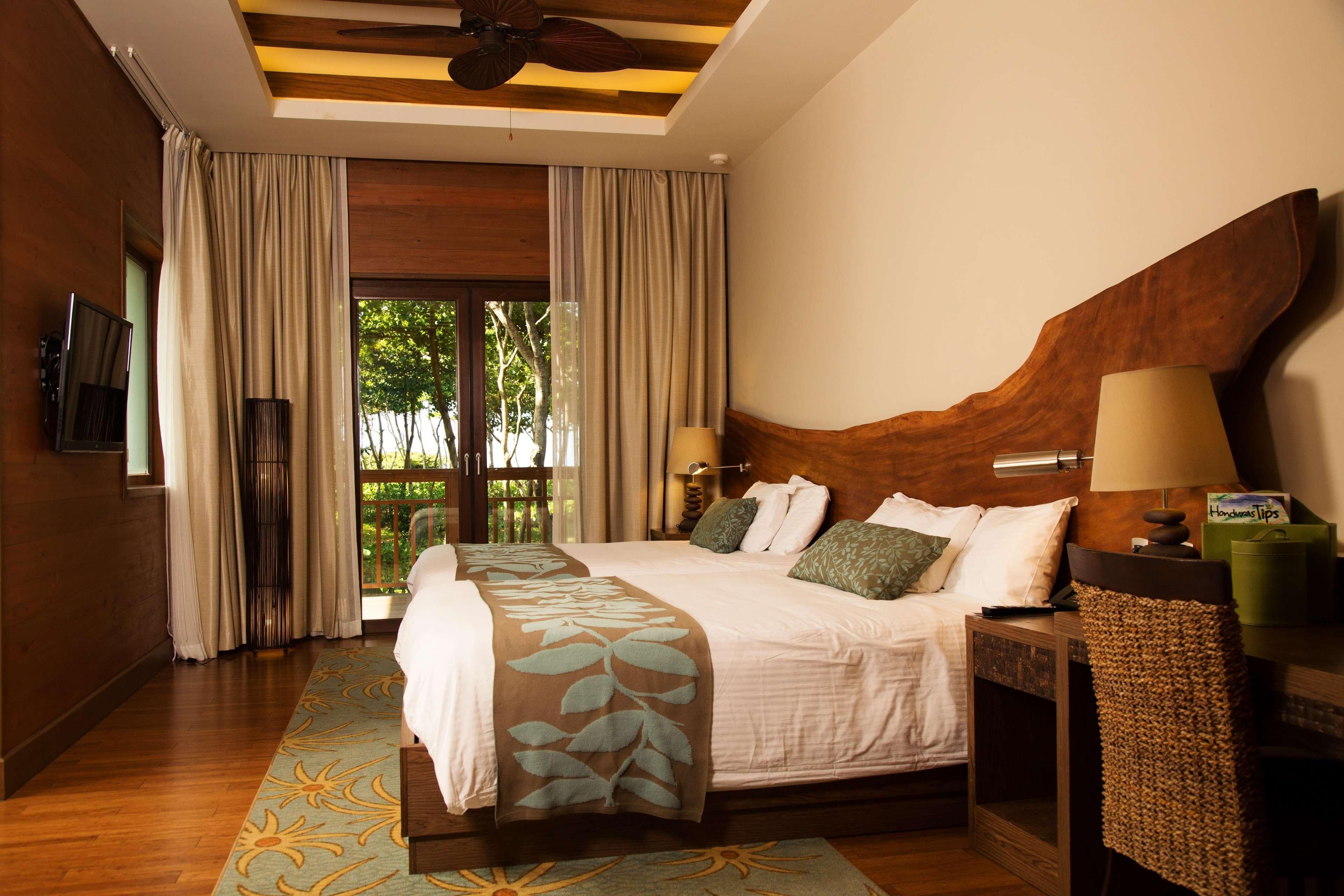 A hotel room with a large bed, a wooden headboard, a TV mounted on the wall, and a view of greenery outside.