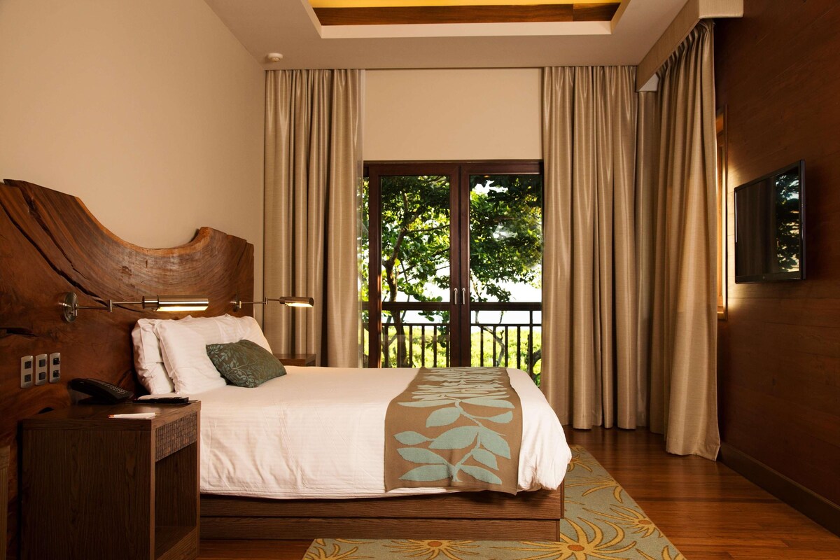 Quarto com cabeceira de madeira, cama com roupa de cama branca, mesa de cabeceira com telefone e controle remoto, e uma janela grande com cortinas.