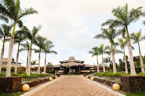 A pathway leading to a building with a thatched roof, flanked by palm trees and a brick walkway.