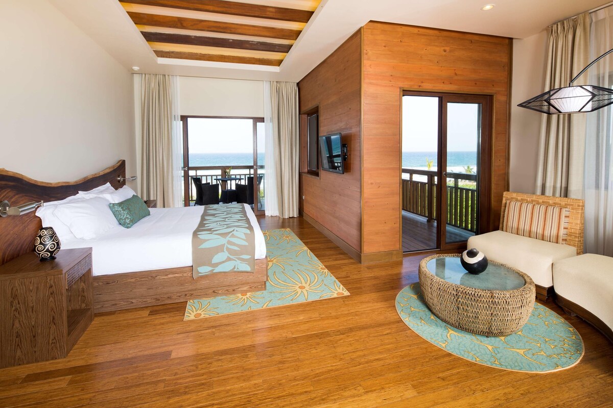 Quarto de hotel moderno com uma cama grande, um sofá, uma TV e uma varanda com vista para o mar.