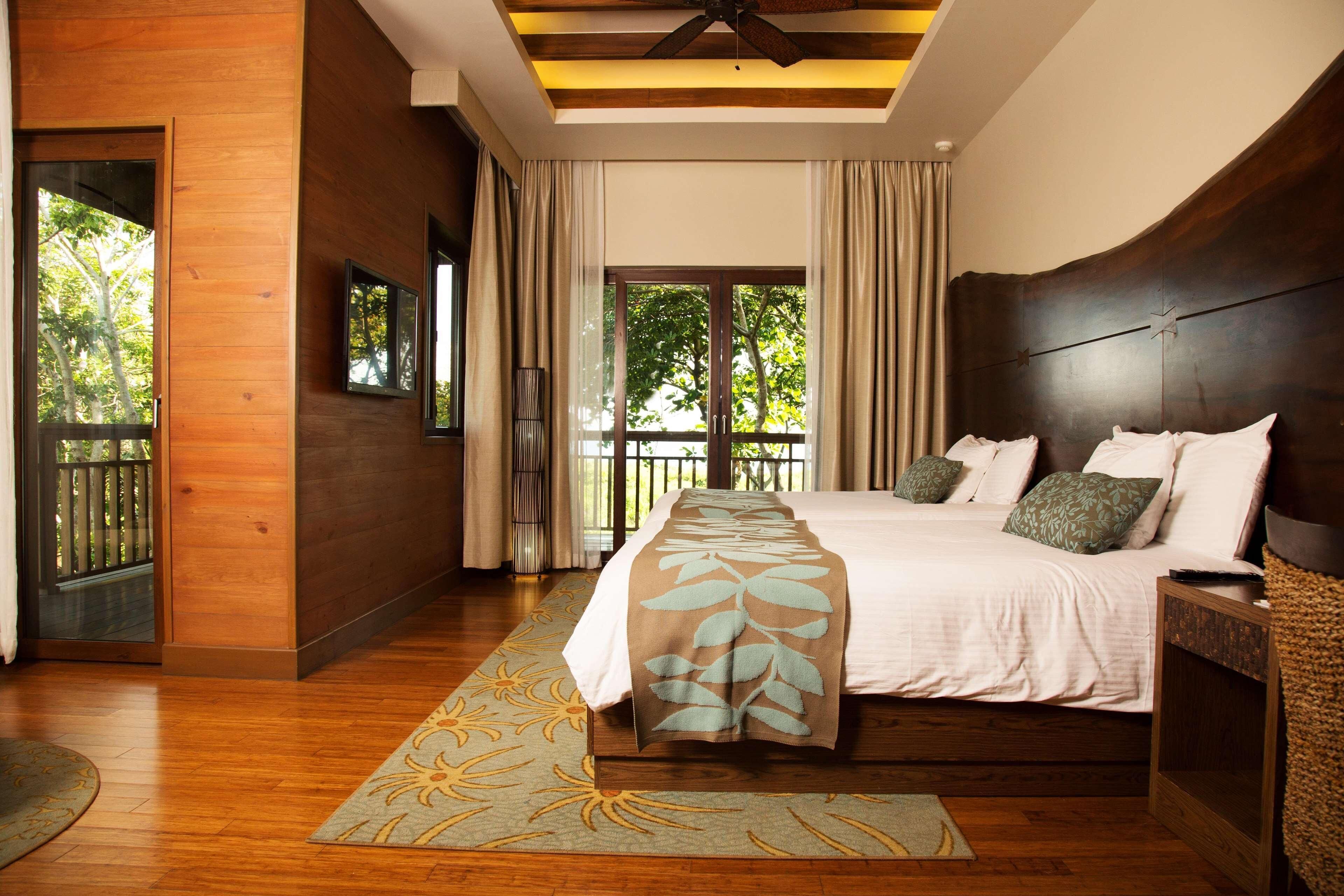A bedroom with a large bed, wooden flooring, a ceiling fan, and a view of the outdoors through a sliding door.