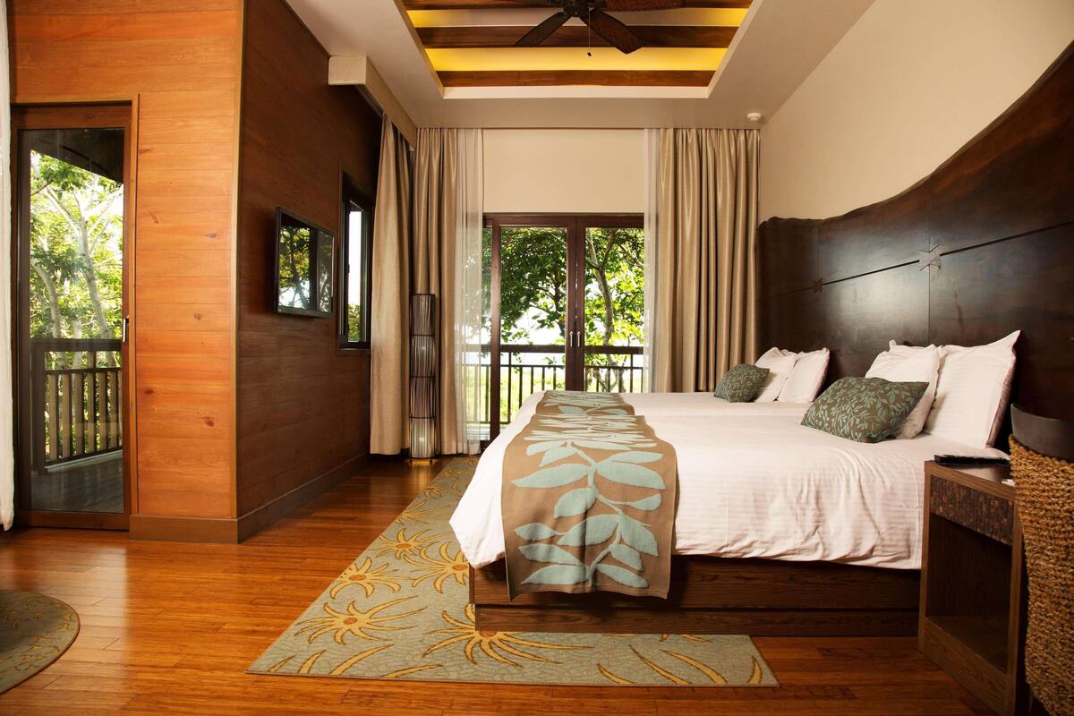 Un dormitorio con una cama grande, piso de madera, ventilador de techo y vistas al exterior a través de una puerta corredera.