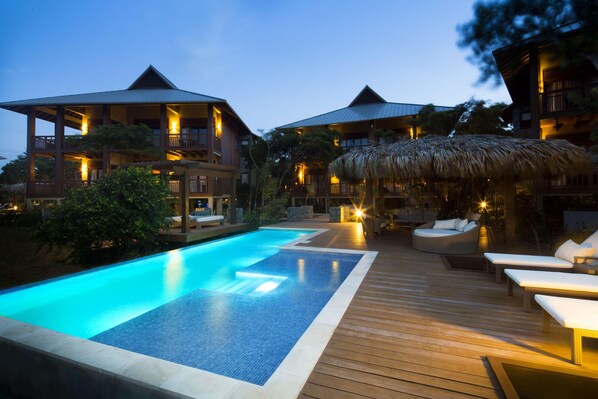 A poolside area with a wooden deck, lounge chairs, and a thatched-roof structure.