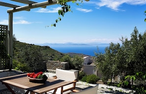 A wooden table with a bowl of fruit, a white cushioned chair, and a view of the sea and hills. - Kea Village (Kea)
