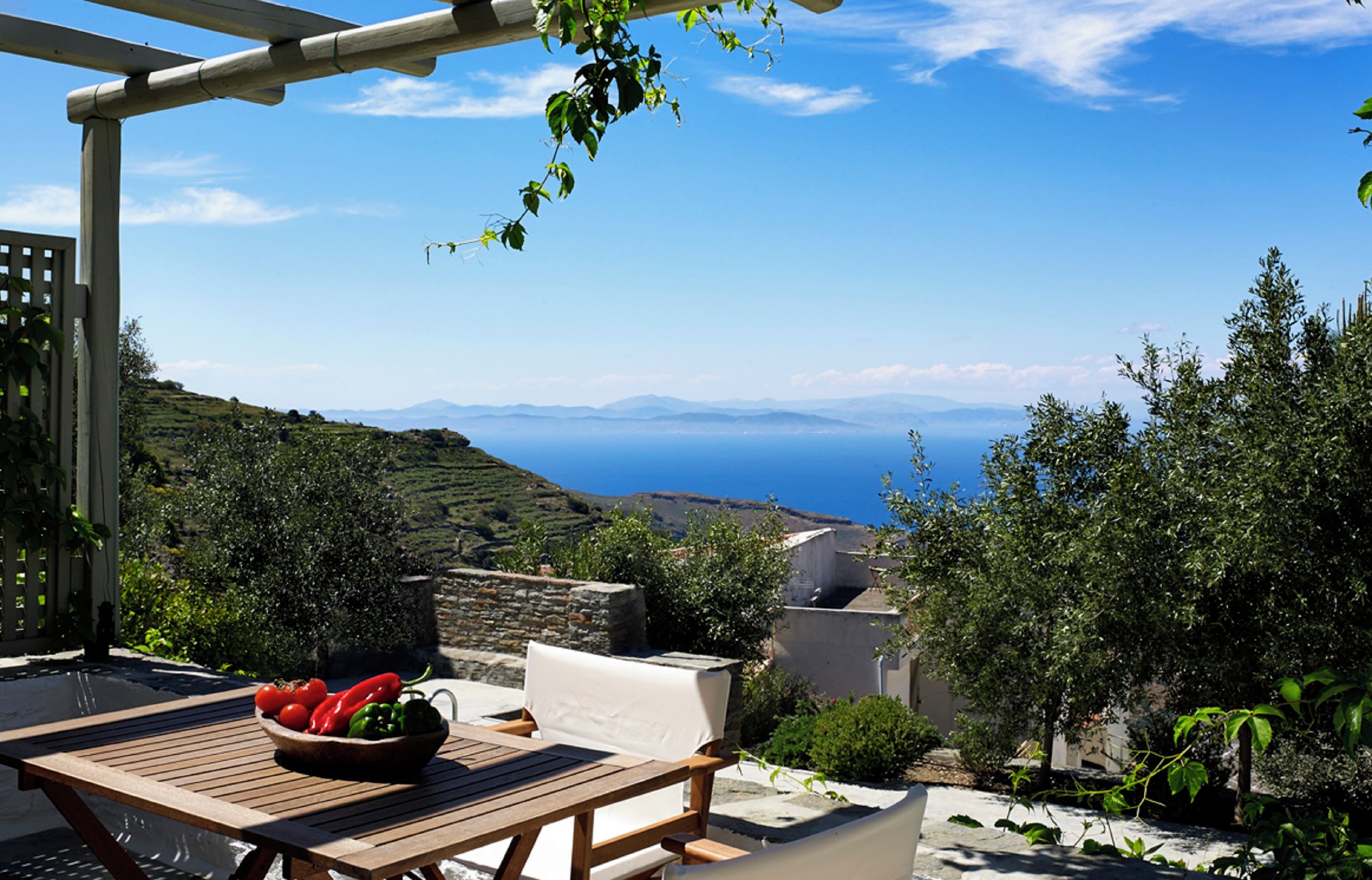 A wooden table with a bowl of fruit, a white cushioned chair, and a view of the sea and hills.