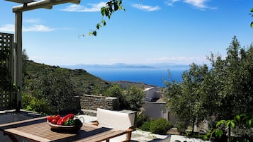 A wooden table with a bowl of fruit, a white cushioned chair, and a view of the sea and hills.
