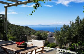 Une table en bois sur laquelle se trouve un saladier rempli de fruits, une chaise blanche rembourrée et une vue sur la mer et les collines.