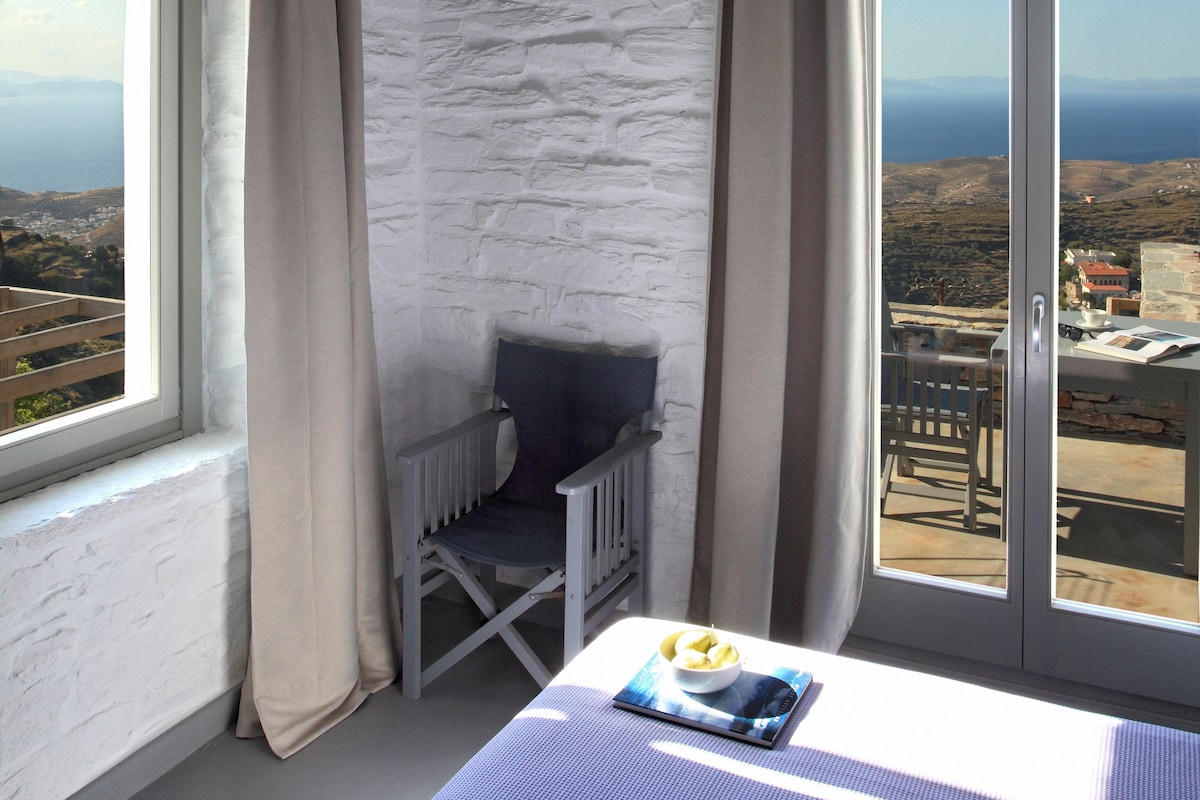 Une chambre avec un lit, une petite table sur laquelle se trouve un saladier de fruits, une chaise et une vue sur la mer par la fenêtre.