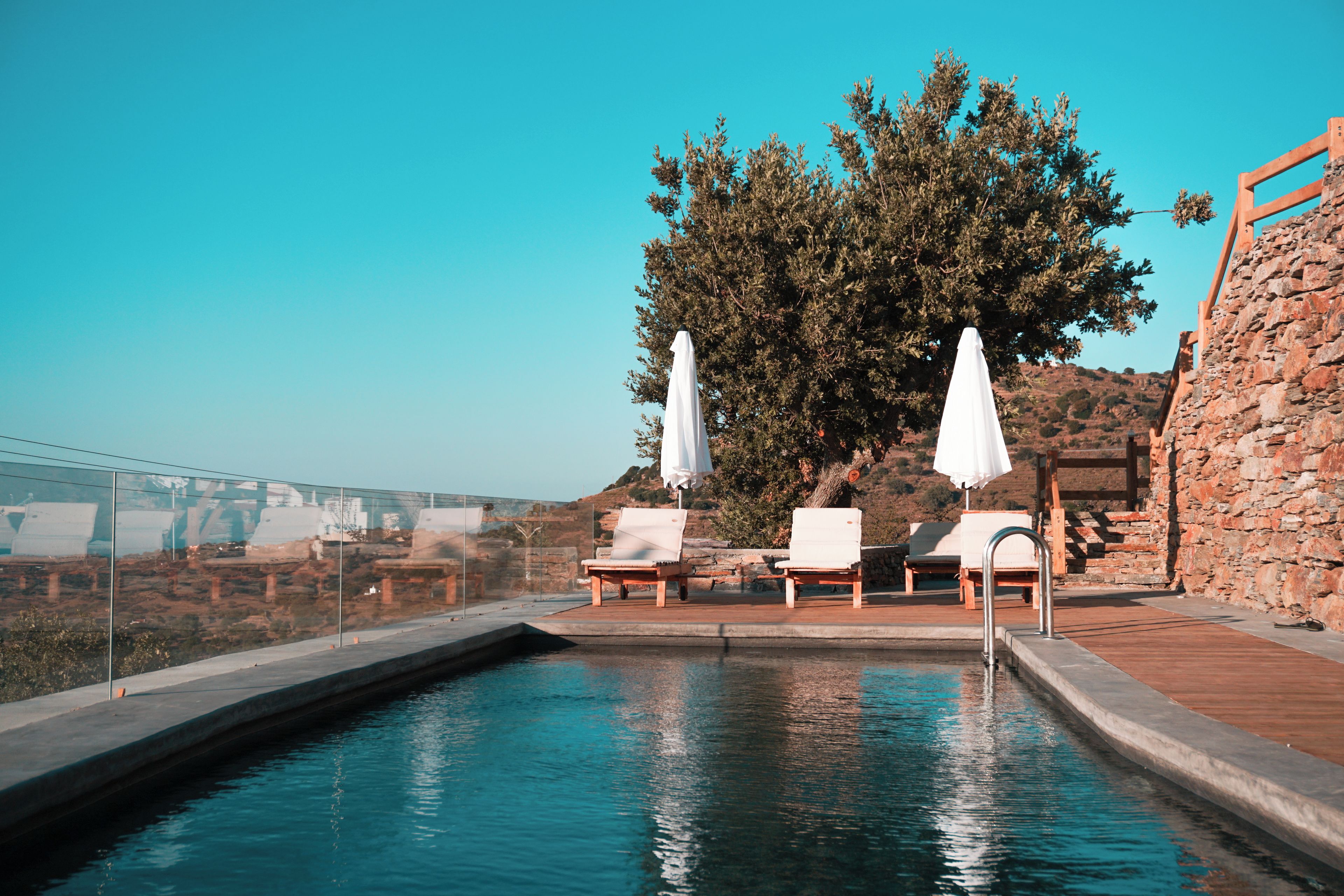 A pool with lounge chairs and umbrellas, surrounded by stone walls and a large tree.