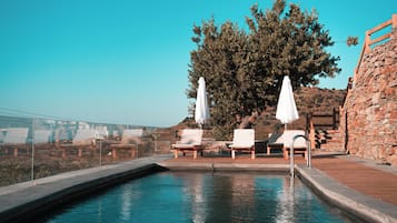 A pool with lounge chairs and umbrellas, surrounded by stone walls and a large tree.