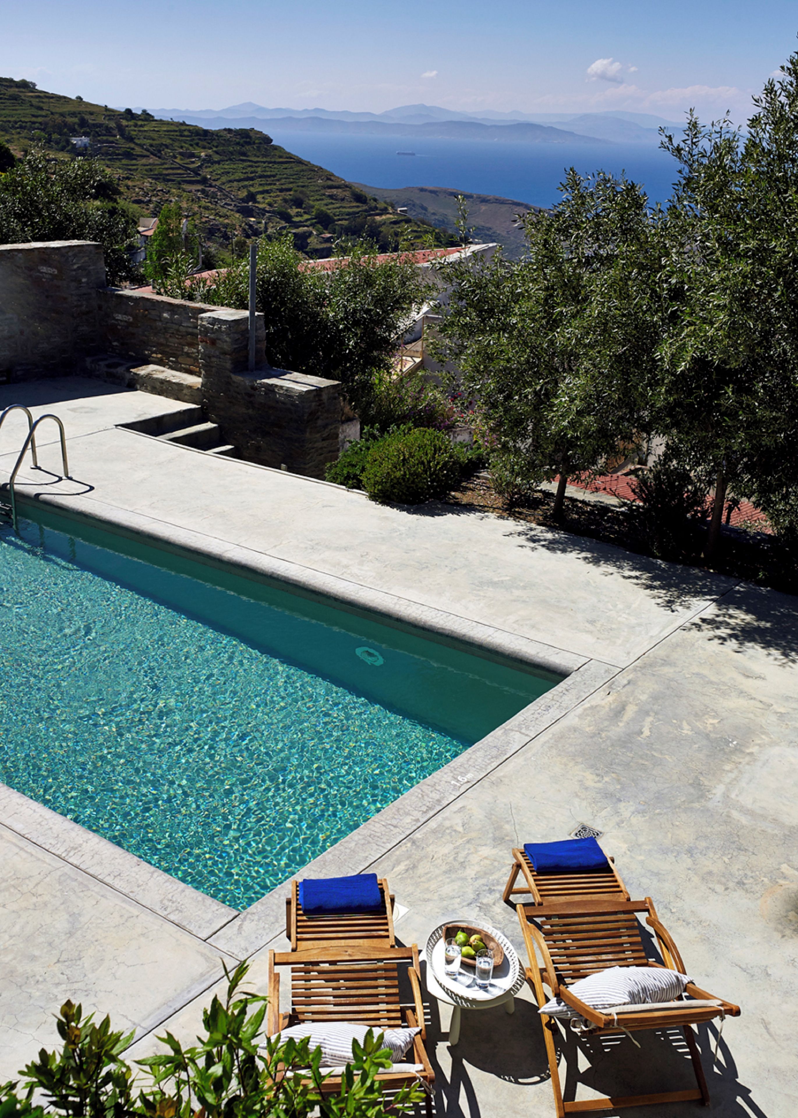 A swimming pool with clear water, wooden lounge chairs, and a view of the sea and hills.