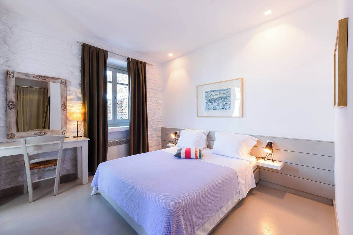 Une chambre d’hôtel moderne comprenant un lit, des lampes de chevet, un bureau avec une chaise, une fenêtre avec des rideaux et un tableau encadré au mur.
