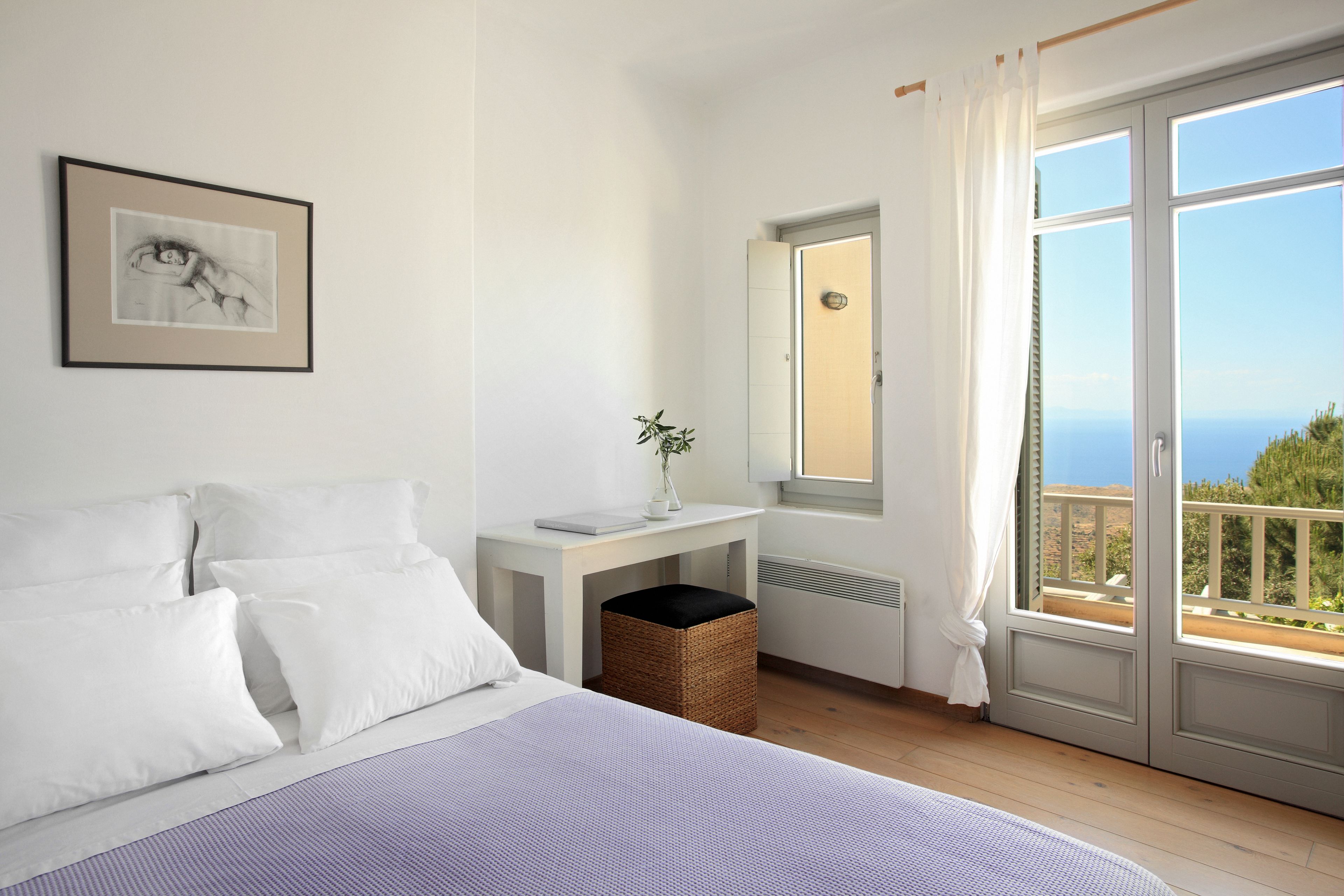 Ein Schlafzimmer mit Bett, Nachttisch mit Vase, Fenster mit Blick und einem gerahmten Bild an der Wand.
