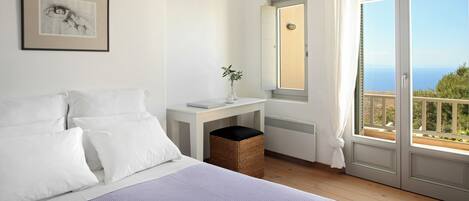 A bedroom with a bed, a bedside table with a vase, a window with a view, and a framed picture on the wall.