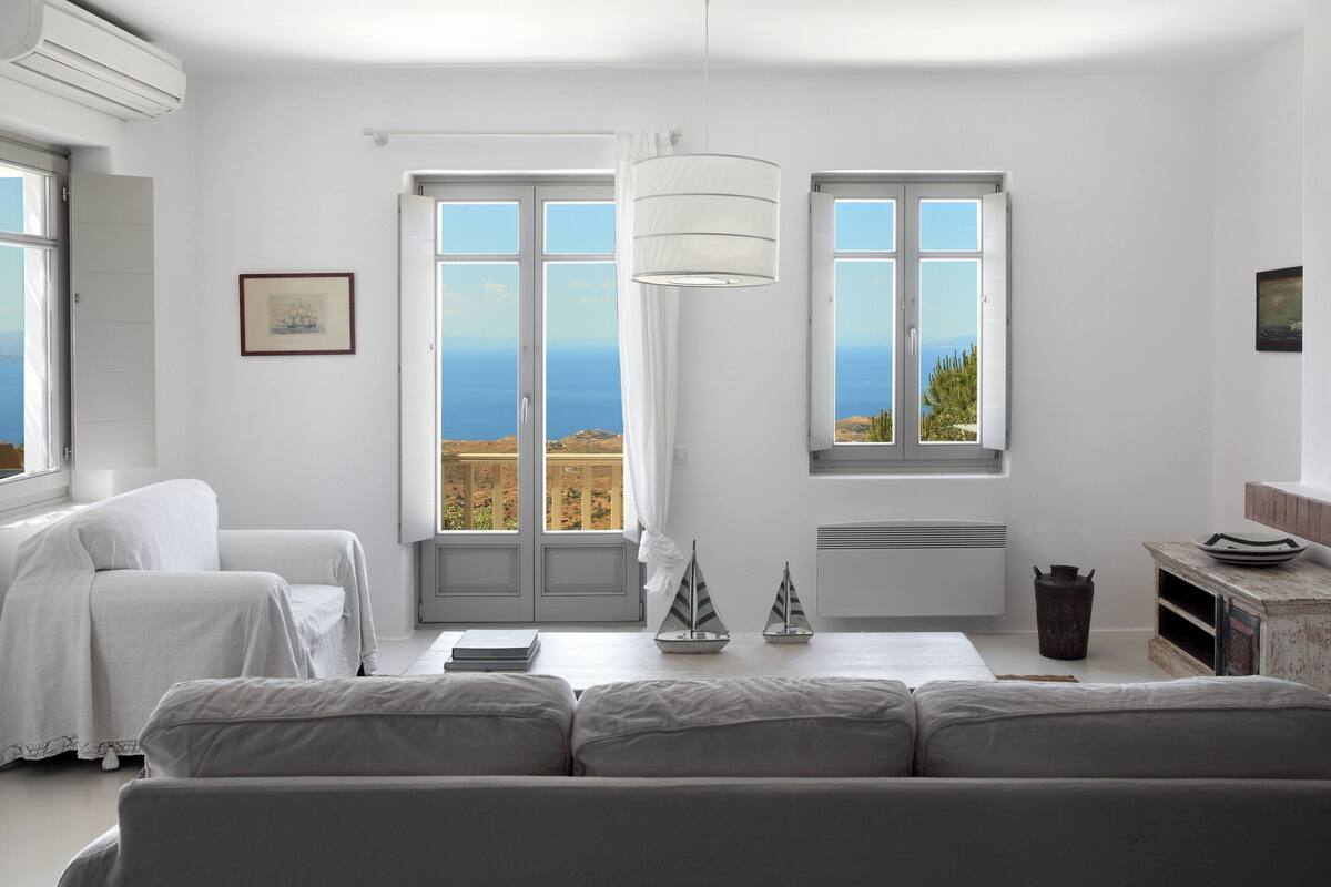 Une pièce au décor minimaliste, avec un canapé blanc, une petite table agrémentée d’objets décoratifs et une vue sur la mer grâce à de grandes fenêtres.