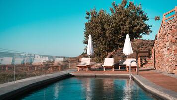 A pool area with lounge chairs and umbrellas, surrounded by stone walls and a large tree.