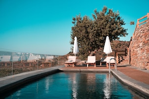 Zona de piscina con sillones de descanso y sombrillas, rodeada de muros de piedra y un árbol grande.