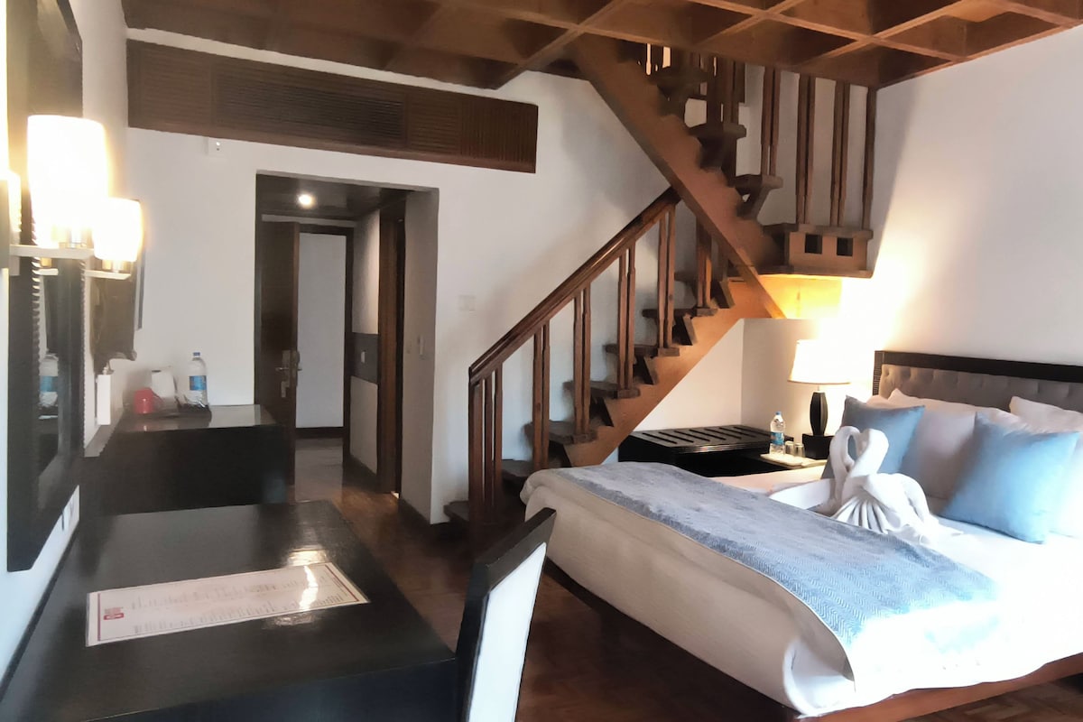 Una habitación de hotel con una cama, un escritorio, una silla, una escalera de madera y una mesita de noche con una lámpara.