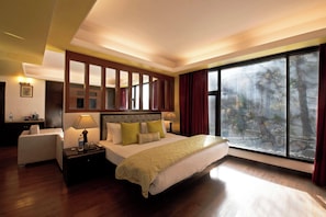 Una habitación de hotel con una cama grande, una mesita de noche, una silla, un ventanal con vistas a árboles e iluminación empotrada en el techo.