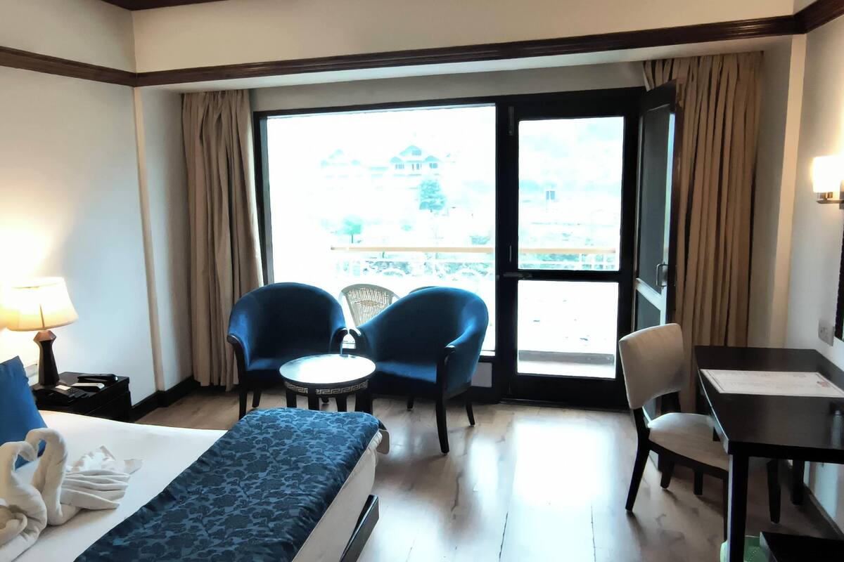 Une chambre d’hôtel avec un lit, deux chaises, une petite table, un bureau et une vue sur l’extérieur grâce à une grande fenêtre.