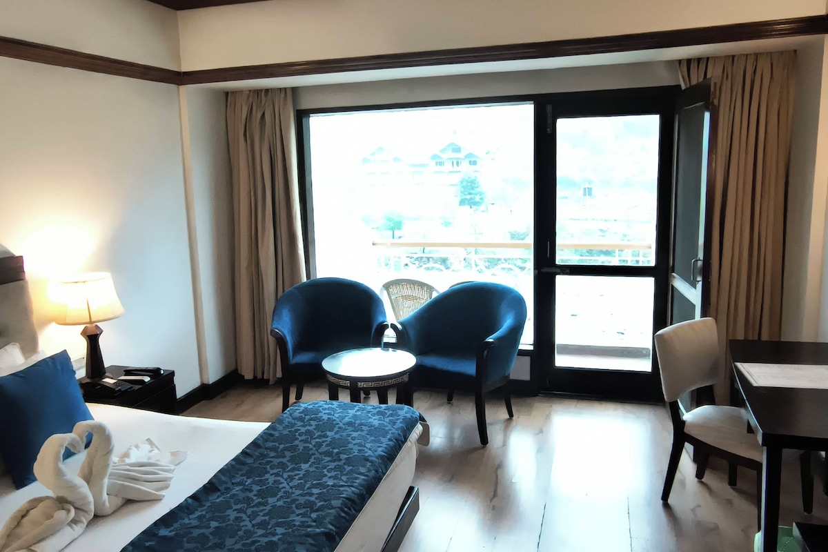 Habitación de hotel con una cama, dos sillas, una mesita, un escritorio y vistas por un ventanal grande.