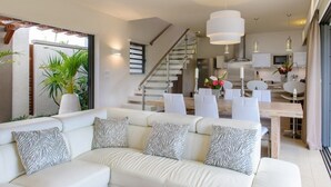 Ein modernes Wohnzimmer mit einem weißen Sofa, zebra-gemusterten Kissen, einem Essbereich mit weißen Stühlen, einer Treppe und einer Küche mit weißen Schränken.