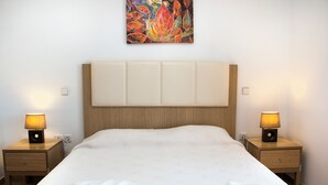 A neatly made bed with a headboard, two bedside tables with lamps, and a colorful painting on the wall. - Gennadi Garden Apartments (Rhodes)