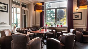 A room with a large window, leather seating, a wooden table, and potted plants. - Hotel La Sapiniere (Hallines)