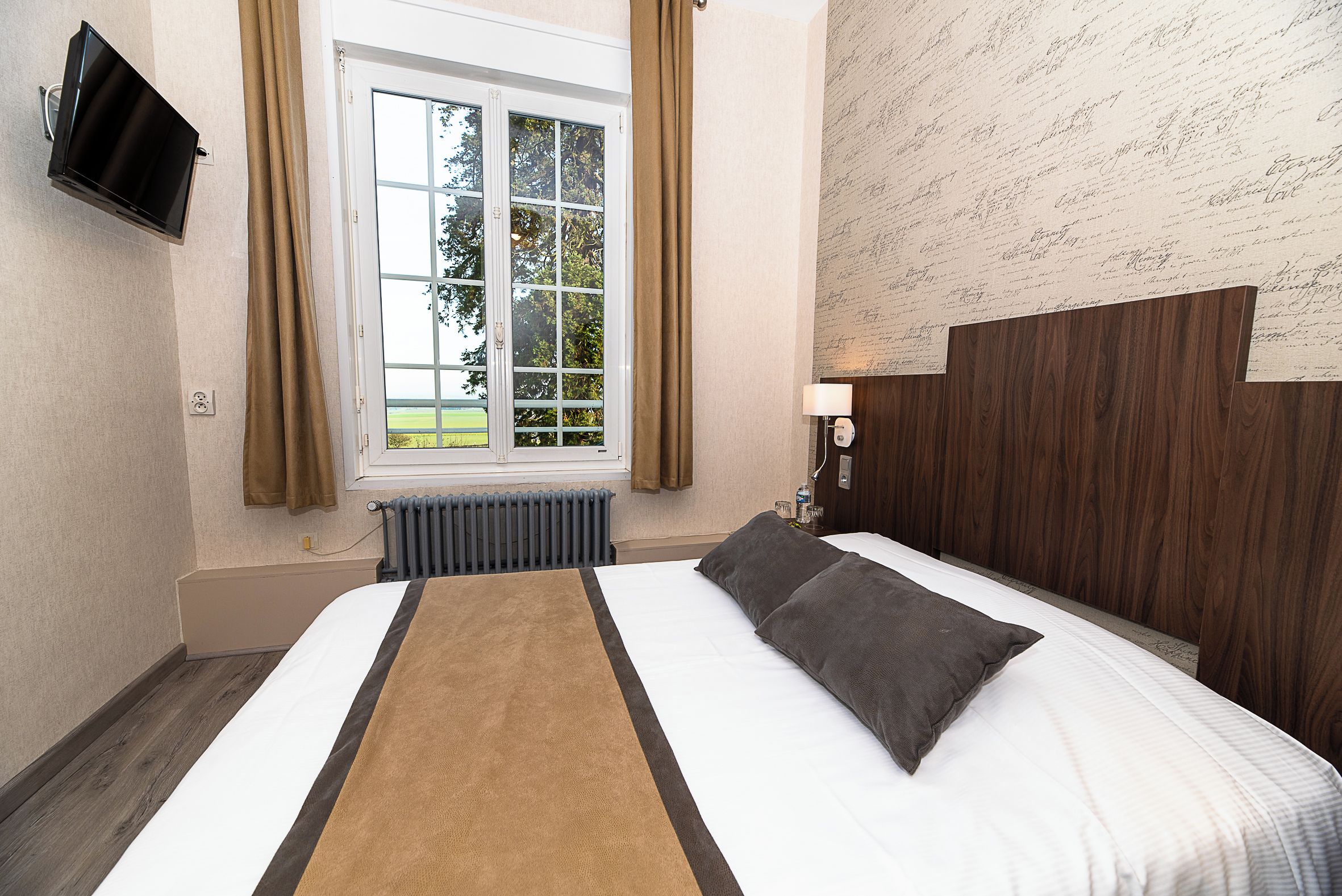 A hotel room with a bed, a window, a radiator, and a television mounted on the wall.