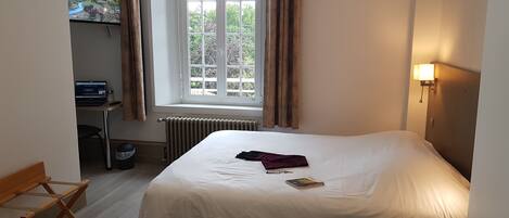 A hotel room with a bed, a desk with a computer, a window with curtains, and a radiator.