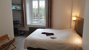 A hotel room with a bed, a desk with a computer, a window with curtains, and a radiator. - Hotel La Sapiniere (Hallines)
