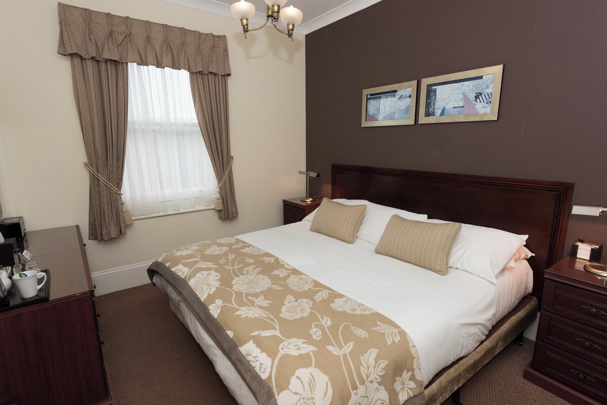 A hotel room with a large bed, a nightstand, a dresser, a window with curtains, and two framed pictures on the wall.