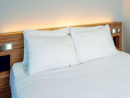 Una cama bien hecha con sábanas y almohadas blancas, cabecera de madera y controles a los lados.