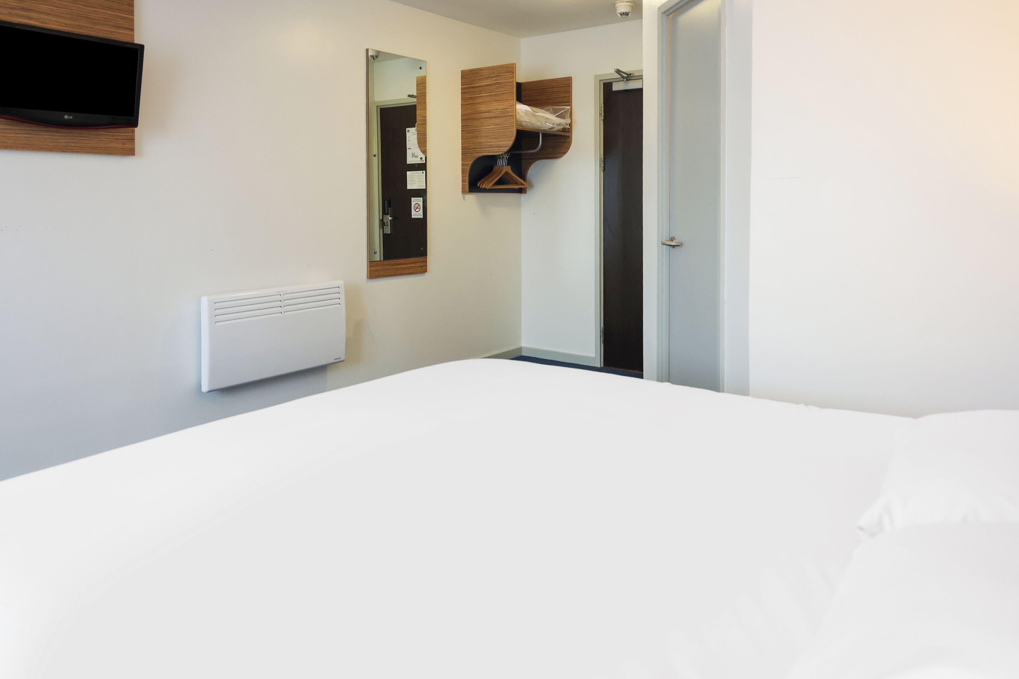 Un dormitorio con una cama, un televisor montado, un espejo, un perchero y una puerta.
