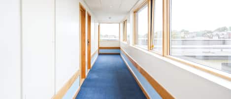 A corridor with a blue carpet, wooden doors, and large windows offering a view of buildings and a bridge.