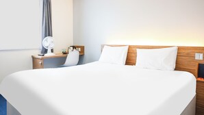 A hotel room with a large bed, a bedside table with a lamp, a chair, and a wall-mounted fan. - Ibis Budget Lancaster Centre (Lancaster)
