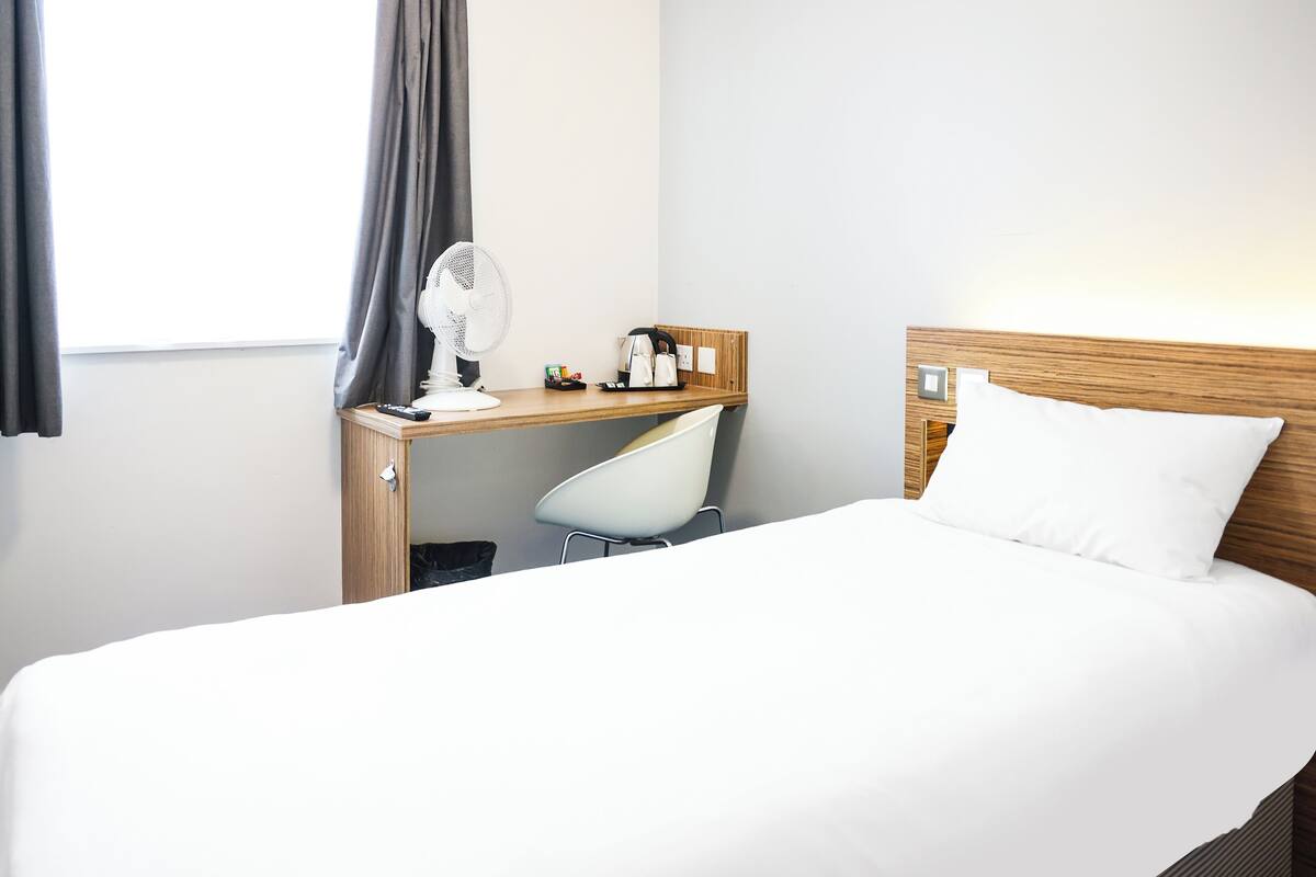 A hotel room with a bed, a desk with a chair, a fan, and a window with curtains.