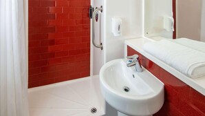 A bathroom with a red brick wall, a white sink, a mirror, and a shower area. - Ibis Budget Lancaster Centre (Lancaster)