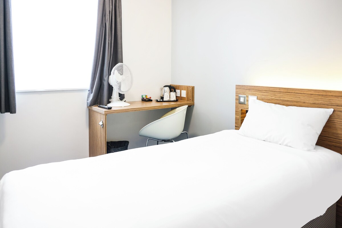 A hotel room with a bed, a desk with a chair, a fan, and a window with curtains.