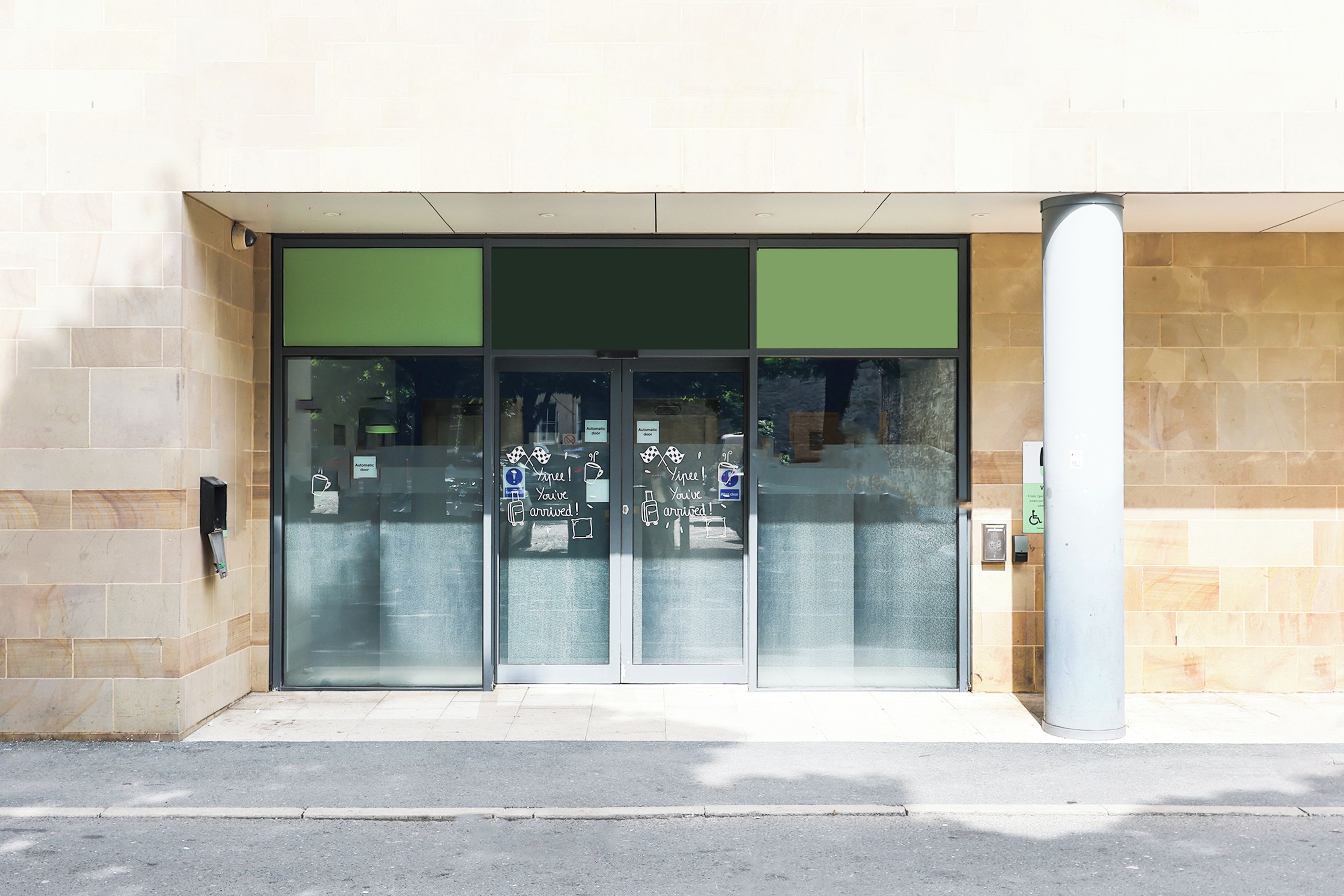Entrada de un edificio con puertas de vidrio, un toldo verde y señal de accesibilidad para sillas de ruedas.