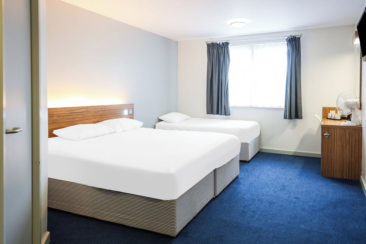 A hotel room with two beds, a window with curtains, a bedside table with a lamp, and a small wooden cabinet.