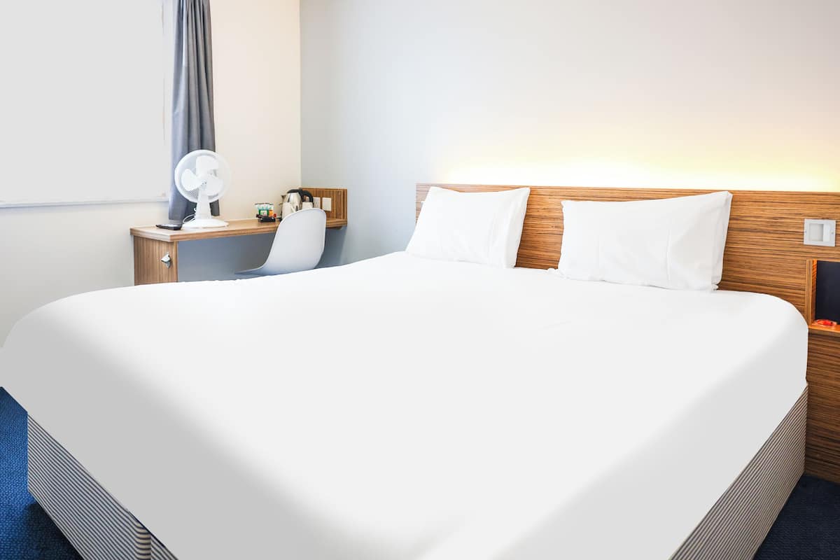 A hotel room with a large bed, a bedside table with a lamp, a chair, and a wall-mounted fan.