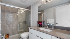 Ein modernes Badezimmer mit Duschkabine aus Glas, einer weißen Toilette, einem Waschbecken inklusive Spiegel und Pflegeprodukten auf der Ablagefläche.