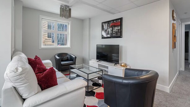 Ein modernes Wohnzimmer mit einem weißen Sofa, einem schwarzen Ledersessel, einem Glastisch, einem Flachbildfernseher und einem Stadtbildposter an der Wand.