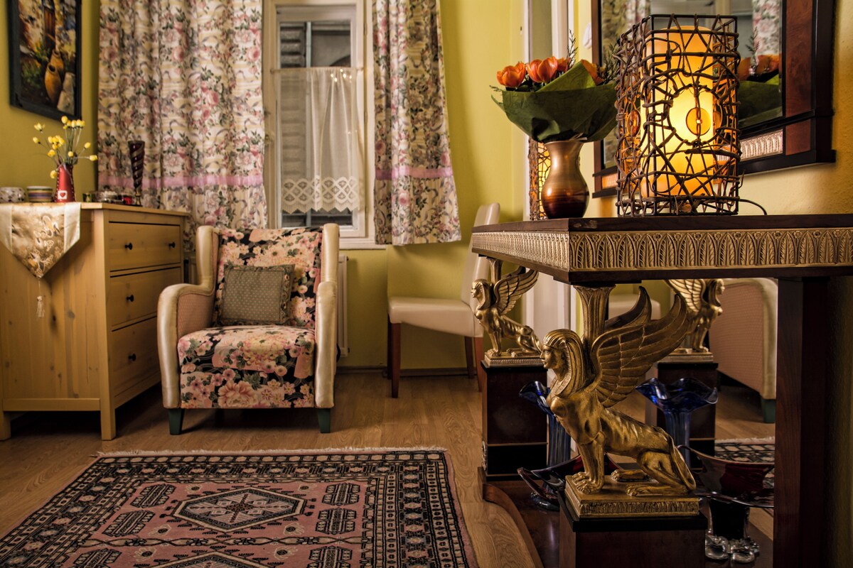 Ein gemütliches Zimmer mit einem blumigen Sessel, einer Holzkommode, einem gemusterten Teppich und Dekorationselementen, darunter eine Vase mit Blumen und eine goldene Statue.