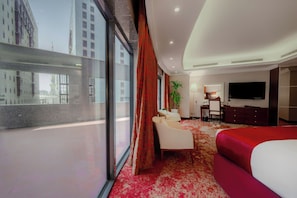 A hotel room with a large window, a bed, a desk, a chair, a TV, and a plant.