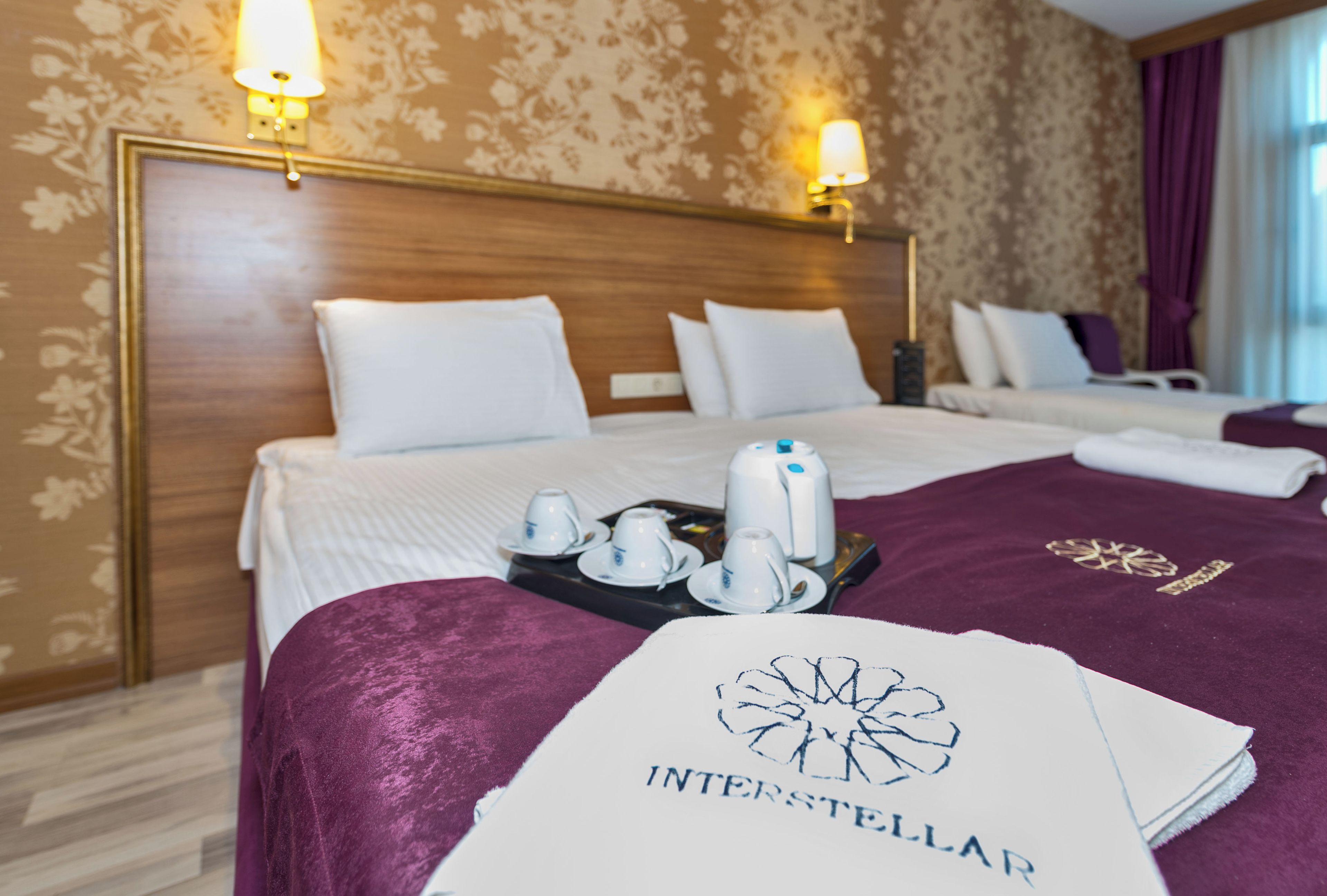 A hotel room with a large bed, two bedside tables, a tray with tea cups, and a towel with 'INTERSTELLAR' branding.
