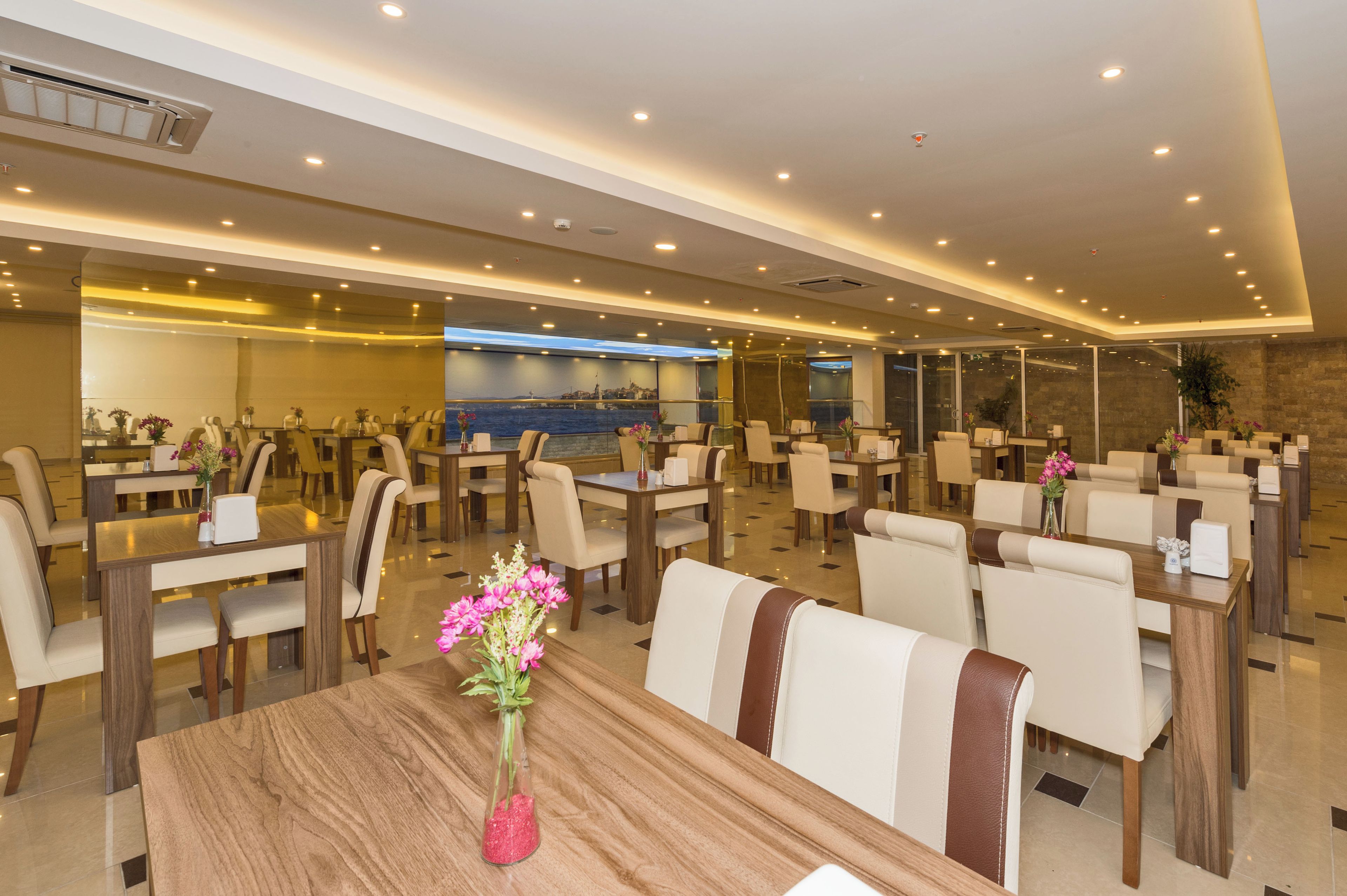 A hotel dining area with multiple tables and chairs, a large wall-mounted screen, and a modern ceiling design.