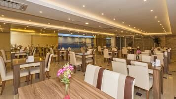 A hotel dining area with multiple tables and chairs, a large wall-mounted screen, and a modern ceiling design.