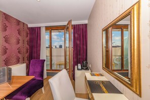 A hotel room with a purple chair, a white chair, a wooden table, a small white table, a large mirror, and a view of the city through a glass door. - Harmony Hotel (Istanbul)